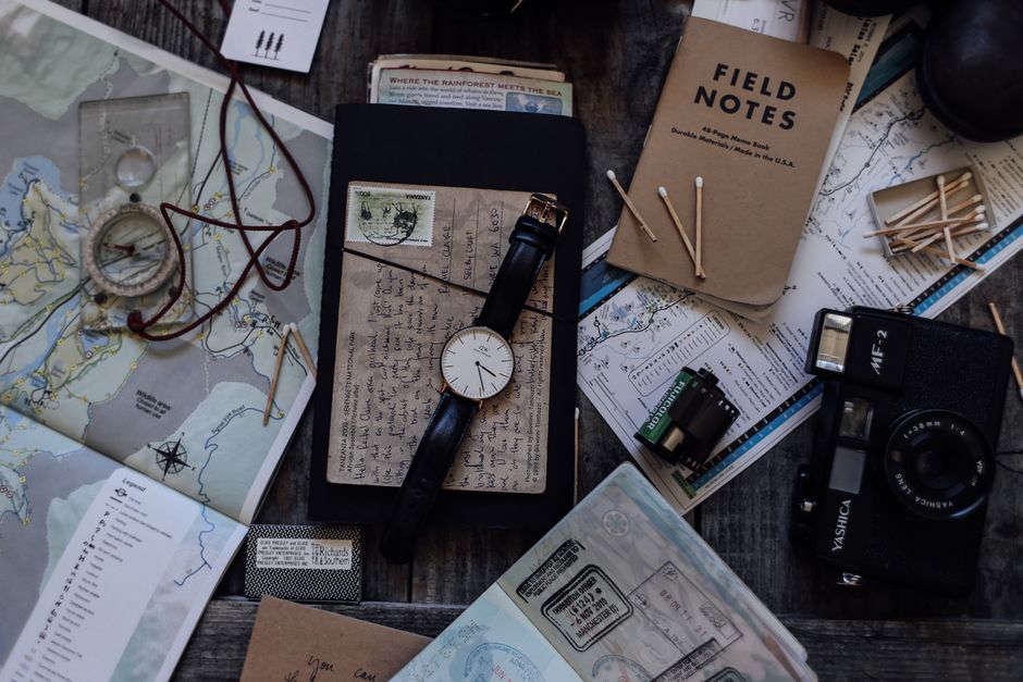 Top view of vintage photo camera near wristwatch and camera roll near maps and notebook near compass and papers near matches placed on table