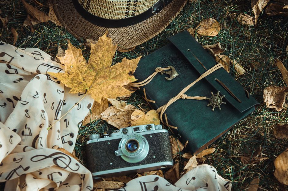 Flat lay of vintage camera, notebook, hat, and autumn leaves on grass.